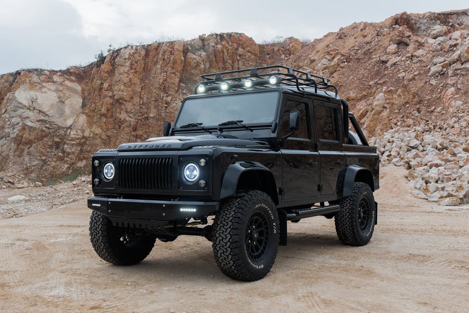 Front View: Custom Land Rover Defender Black custom Land Rover Defender with off-road tires and roof rack in rugged terrain