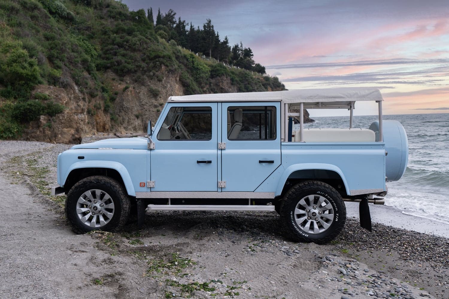 Side View: Bespoke Land Rover Defender A custom Land Rover Defender in light blue parked on a beach with a sandy shoreline and ocean waves.
